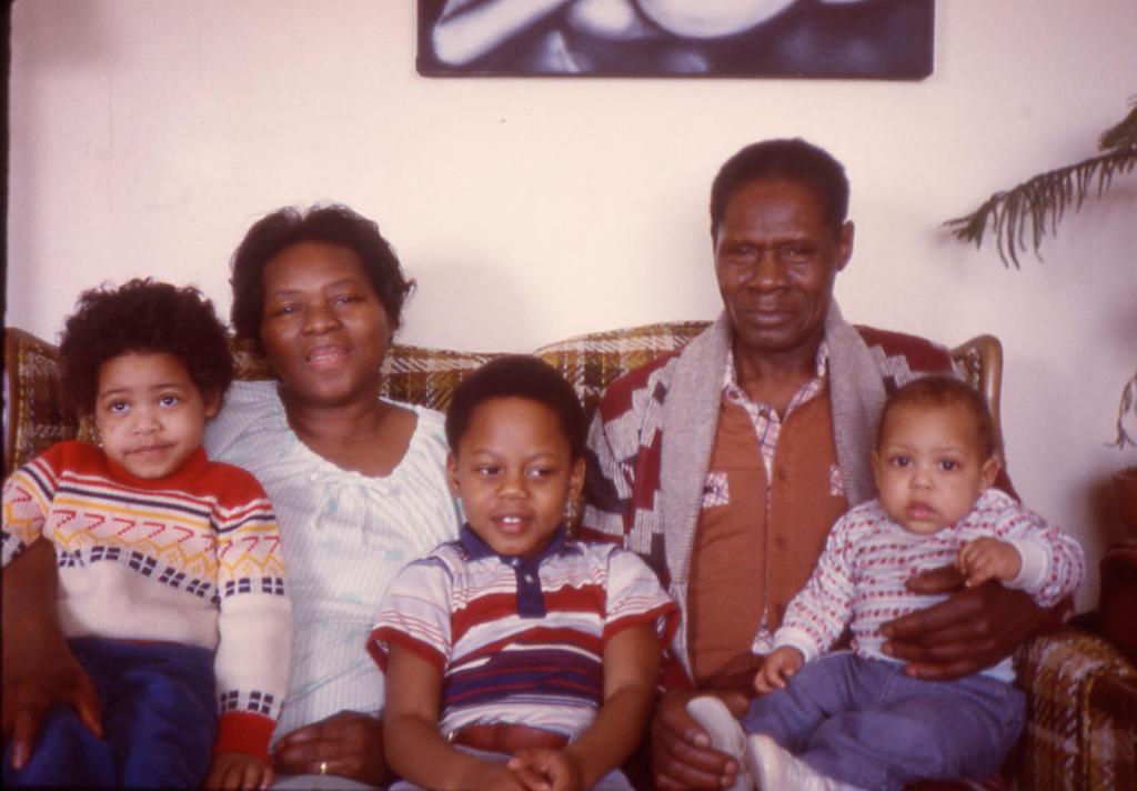 Older Black Couple sitting on a couch with 3 grandchildren--toddler, grade school age, and baby around 1979-1980.