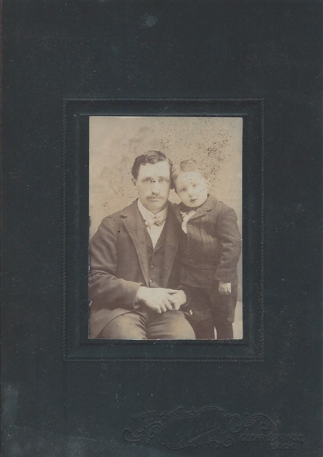 Black matted sepia photograph of Chapin Holden seated and young son Heman standing beside him. Facing the camera with head together.